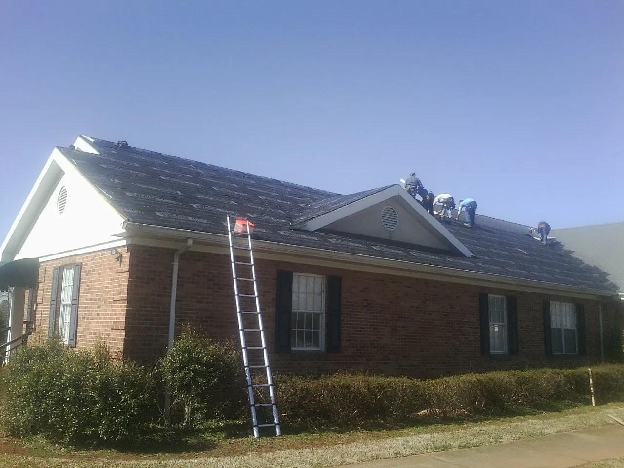 Crew tearing shingles off a brick ranch roof with a ladder set — Emergency Roof Repair work by our crew in Belle Glade