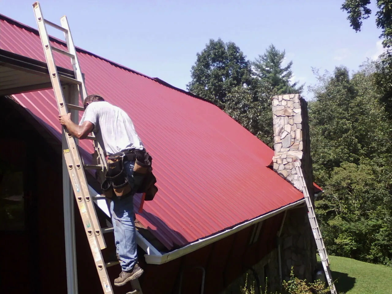 Roofer on a ladder beside a red metal roof and stone chimney — Soffit & Fascia Repair by Roofing Brothers in Belle Glade